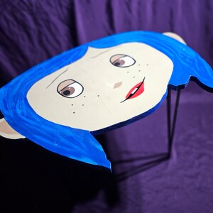 May include: A blue and white wooden table top with a cartoon face of a girl with blue hair and red lips. The table top is supported by a black metal stand.