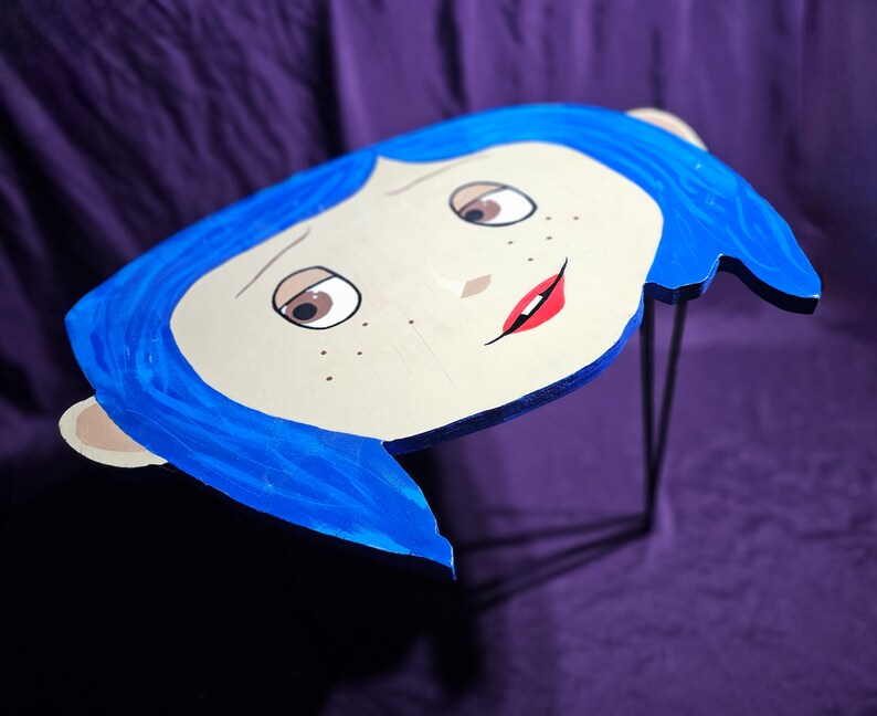 May include: A blue and white wooden table top with a cartoon face of a girl with blue hair and red lips. The table top is supported by a black metal stand.