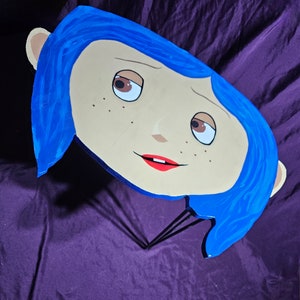 May include: A blue and beige wooden cutout of a girl's face with blue hair, red lips, and brown eyes. The cutout is attached to a black metal stand.