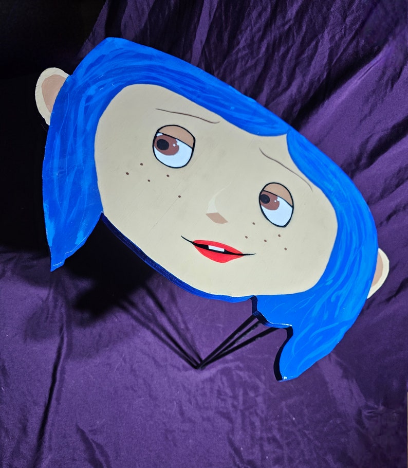 May include: A blue and beige wooden cutout of a girl's face with blue hair, red lips, and brown eyes. The cutout is attached to a black metal stand.