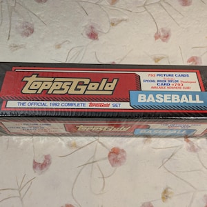 May include: Sealed Topps Gold baseball card set from 1992. The box is black with red and gold accents, and the text "Topps Gold" is prominently displayed. The box includes 793 picture cards and a special Brien Taylor card.