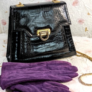May include: A black handbag with a glossy crocodile-embossed texture, featuring a gold-tone metal clasp and a matching gold chain strap, paired with a pair of deep purple suede gloves with stitched detailing on the back of the hands.