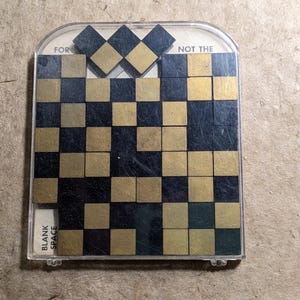 May include: A vintage puzzle game with a clear plastic cover. The game features a grid of alternating black and gold squares, with some squares missing. The text "FOR" and "NOT THE" are visible on the top, and "BLANK SPACE" on the bottom.