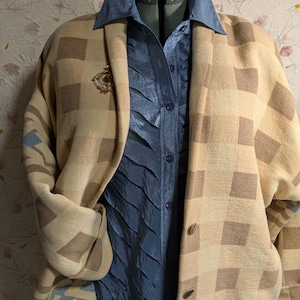 May include: A tan and brown checkered cardigan with a blue shirt underneath. The cardigan has a geometric pattern and a small gold brooch. The blue shirt has a textured design and a button-up front. The outfit is displayed on a dress form.