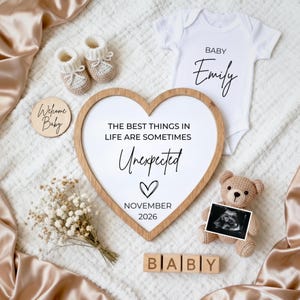 Pode incluir: Uma composição para um anúncio de gravidez com uma moldura de madeira em forma de coração e o texto "THE BEST THINGS IN LIFE ARE SOMETIMES Unexpected". Inclui um body, uma ecografia, botinhas e um ursinho de peluche.