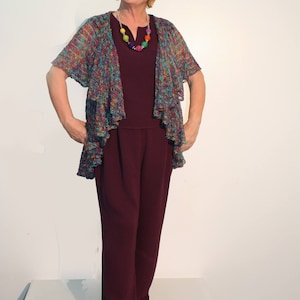 Op de afbeelding: Een vrouw draagt een bordeauxrode jumpsuit met een kleurrijk gehaakt vest en een kralenketting. Ze draagt bruine schoenen.