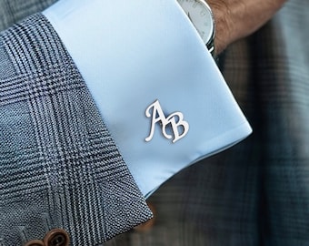May include: A man wearing a gray plaid suit jacket with a light blue shirt and a light blue patterned tie. He is wearing cufflinks with the initials "AB" on them.