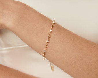 Op de afbeelding: Een gouden kettingarmband met kleine witte parels. De armband heeft een delicate, vrouwelijke uitstraling.