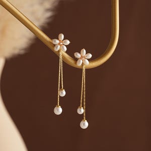 Puede incluir: Elegantes pendientes dorados con diseño floral y detalles de perlas. Cada pendiente tiene una flor con cadenas colgantes y tres perlas blancas. Los pendientes se exhiben en un expositor de joyería dorado.