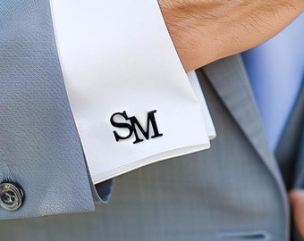 May include: A gray suit jacket with a white shirt and a blue tie. The shirt has black cufflinks with the initials "SM" embroidered on them.