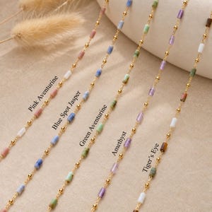 May include: Five delicate gold-toned chains featuring cylindrical gemstone beads in various colours. The labels identify the stones as Pink Aventurine, Blue Spot Jasper, Green Aventurine, Amethyst, and Tiger's Eye, interspersed with small gold-toned metal spheres.