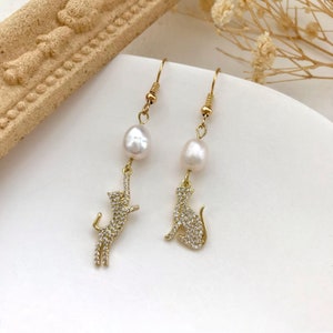 May include: Gold dangle earrings with a pearl and a cat charm. The cat charm is covered in small clear rhinestones.