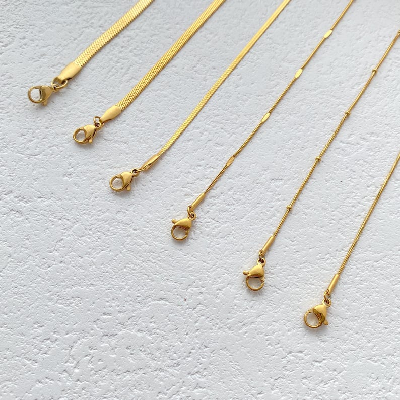 May include: Five gold chains of different styles and lengths, all with lobster claw clasps. The chains are laid out on a white textured surface.