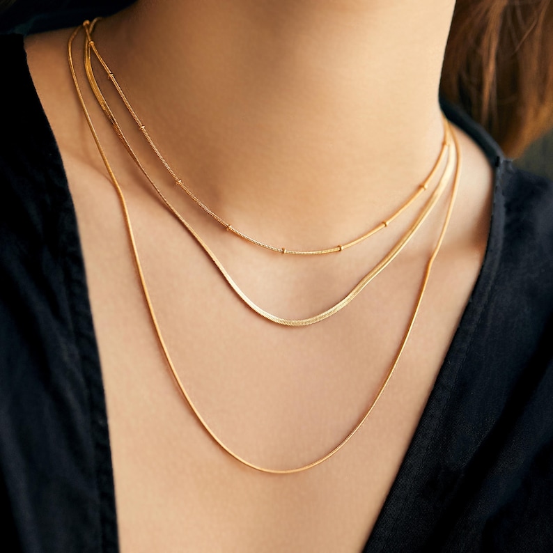 May include: Three gold chain necklaces layered on a person's neck. The chains are different widths and styles.