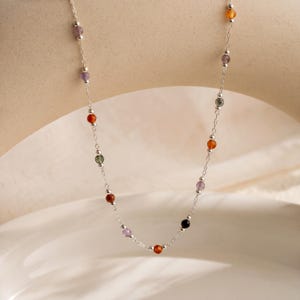 May include: A delicate silver chain necklace featuring small, evenly spaced, multi-colored gemstone beads. The necklace includes round beads in shades of orange, purple, green, and black, interspersed with tiny silver metal spacer beads.