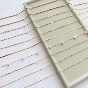 May include: A collection of gold chain necklaces of varying styles and lengths, displayed on a light beige surface. Some chains feature small pearl accents.