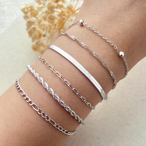 Puede incluir: Una colección de pulseras de plata de varios estilos, incluyendo una cadena de espiga y una cadena de cuerda. Las pulseras se muestran en una muñeca.