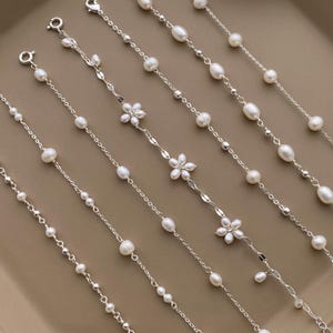 Puede incluir: Un conjunto de seis delicados brazaletes de cadena de plata con detalles de perlas. Algunos brazaletes presentan pequeños abalorios con forma de flor.