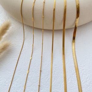 May include: Five gold chains of different styles and widths, laid out on a white surface.