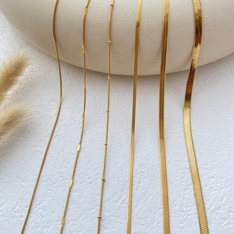 May include: Five gold chains of different styles and widths, laid out on a white surface.