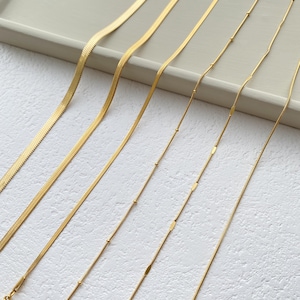 May include: Six gold chains of different styles and lengths laid out on a white surface. The chains are all made of a thin, delicate metal.