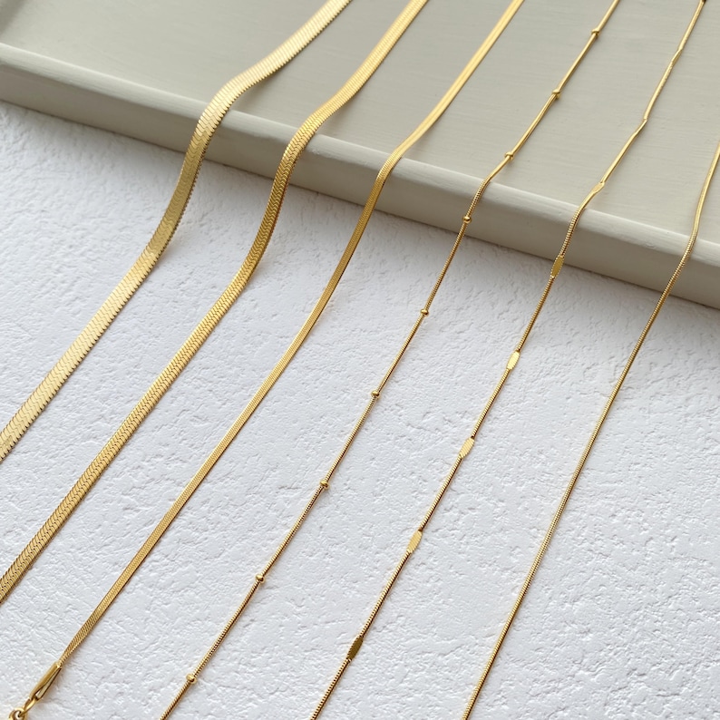 May include: Six gold chains of different styles and lengths laid out on a white surface. The chains are all made of a thin, delicate metal.