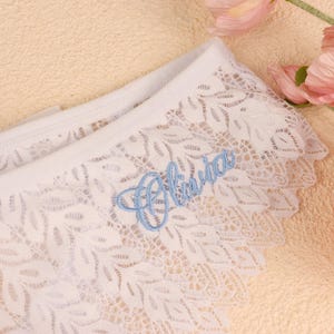 May include: White lace underwear featuring a scalloped edge and a delicate leaf pattern throughout the fabric. The garment is personalised with the name Olivia embroidered in light blue script thread on the front right side of the waistband.