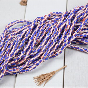 May include: Strands of colorful beads in blue, red, and white. The beads are rectangular and strung together, creating a vibrant pattern. A small, decorative broom is in the foreground, adding a rustic touch. The beads are likely for jewelry making.