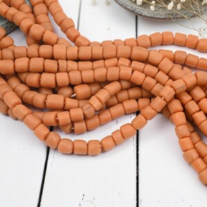 May include: A collection of multiple strands of matte, terracotta-colored beads. Each bead features a rounded, square-like shape and is strung together on a thin cord. The beads have a uniform, earthy orange hue and a smooth, non-reflective surface texture.