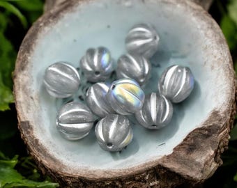 May include: A collection of gray, ridged beads with iridescent highlights, resting in a light blue, rustic bowl. The beads have a spherical shape with vertical grooves and a subtle sheen. The bowl is set on a wooden surface.