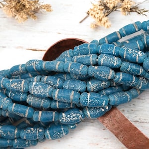 May include: Strands of blue barrel-shaped beads with white accents. The beads have a distressed, aged appearance. The beads are arranged in a pile, with a copper-colored scoop in the background. Dried flowers are in the background.