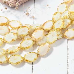 May include: Strands of oval-shaped beads with a creamy white center and a golden-brown edge. The beads are arranged on a white wooden surface, with a few dried floral accents in the background. These beads are ideal for jewelry making.