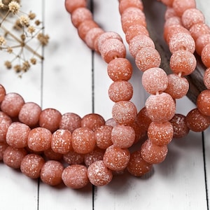 May include: Strands of coral-colored beads with a textured, speckled surface. The beads are round and strung together, displayed on a white wooden surface. The beads are a warm, earthy tone, suggesting natural materials.