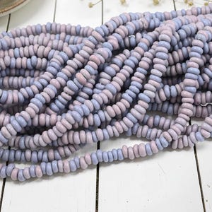 May include: Close-up of a pile of pastel-colored beads. The beads are a mix of light purple and pale pink, arranged in multiple strands. The beads are small, round, and uniform in size, suggesting they are for jewelry making or crafts.
