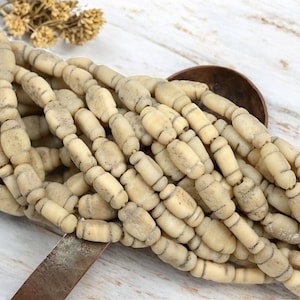 May include: A strand of off-white, barrel-shaped beads with subtle gray markings. The beads are strung together, creating a textured, organic look. The beads are displayed on a light-colored wooden surface.