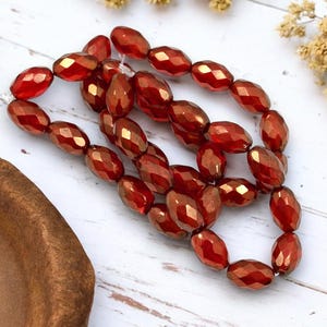 May include: A strand of red faceted glass beads with a metallic sheen. The beads are oval shaped and have a slightly irregular shape.