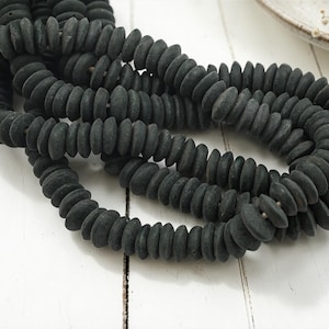 May include: Strands of dark gray, flat, round beads. The beads are strung together, forming multiple loops. The beads are arranged on a white surface, with a small, off-white dish visible in the background. The beads appear to be made of a matte material.