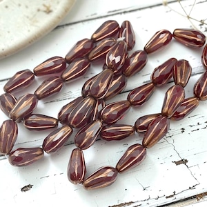 May include: A strand of teardrop-shaped glass beads in a deep burgundy red color. Each bead features a faceted surface with metallic bronze-colored vertical stripes running along the length of the bead, creating a textured and reflective appearance.