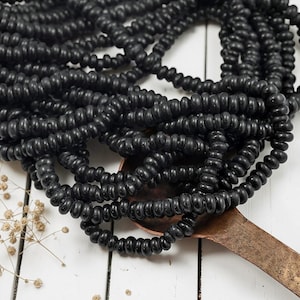 May include: Close-up of multiple strands of black beads. The beads are small, round, and strung together, creating a textured, layered effect. The beads are resting on a copper-colored spoon, with a white wooden surface in the background.