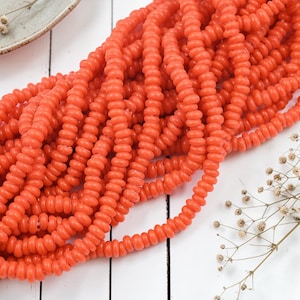 May include: Close-up of a collection of coral-colored beads. The beads are small, round, and strung together, creating a textured, organic look. The beads are arranged on a white wooden surface, with a small ceramic dish and dried flowers in the background.