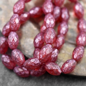 May include: A strand of faceted, oval-shaped beads in a deep red hue. The beads have a textured, almost crystalline appearance, catching the light to create a shimmering effect. The beads are strung together, suggesting their use in jewelry making.