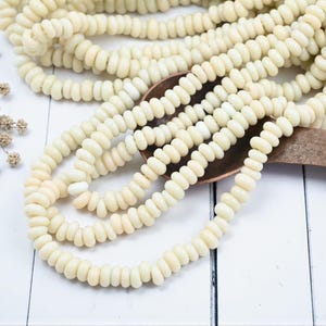 May include: Cream-colored, rounded beads strung together, forming multiple strands. The beads are arranged on a wooden spoon, with some strands spilling onto a white surface. Dried floral accents are visible.