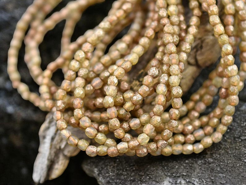 Op de afbeelding: Een streng iriserende gouden glazen kralen met een gefacetteerde, onregelmatige vorm. De kralen zijn geregen op een doorzichtige draad en hebben een glinsterende, metallic afwerking.