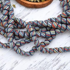 May include: Strands of blue, red, yellow, and white striped beads. The beads are cylindrical and appear to be made of glass or ceramic. The beads are arranged in a curved pattern, suggesting they are part of a necklace or bracelet. The background is a white wooden surface.