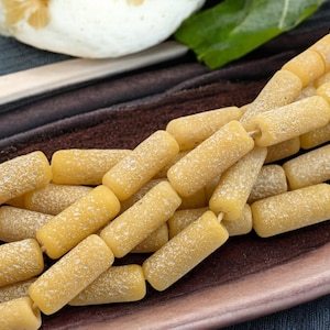 May include: A strand of yellow glass beads with a textured surface. The beads are cylindrical in shape and have a frosted appearance.