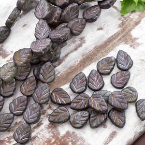 May include: A strand of dark green glass beads in the shape of leaves. The beads have a metallic sheen and are strung on a clear thread. The beads are scattered on a white wooden surface.