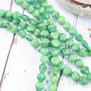 May include: A collection of translucent teal glass beads shaped like small seashells, each featuring delicate yellow vertical stripes. These beads are strung together in multiple strands, showcasing a consistent ribbed texture and a glossy finish.