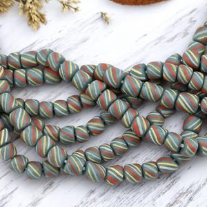 May include: Strands of vintage beads in a muted teal color, each bead adorned with diagonal stripes of red, yellow, and white. The beads are strung together, creating a textured and colorful pattern. Ideal for jewelry making or crafting.