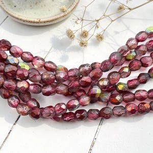 May include: Strands of faceted, oval-shaped beads in shades of burgundy and iridescent hues. The beads are strung together, creating a textured and colorful display. The beads are likely for jewelry making.
