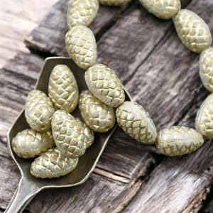 May include: A scoop filled with gold-colored pine cone shaped beads. A strand of the same beads is draped over a piece of weathered wood.